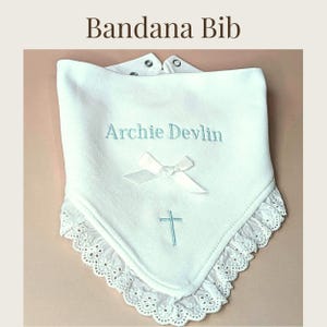 A white bib with the name "Archie Devlin" embroidered on it, along with a small cross symbol, is displayed on a beige background.