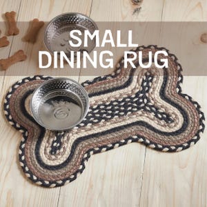 May include: A bone-shaped braided rug featuring concentric patterns in black, tan, and cream, topped with two stainless steel bowls. The text "SMALL DINING RUG" is overlaid on the image. Several bone-shaped treats are visible nearby.