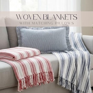 a cozy living space with a gray couch and a striped blanket draped over it. The blanket has a mix of red and white stripes, and there is a matching gray and white striped pillow on the couch. The text overlay reads "WOVEN BLANKETS WITH MATCHING PILLOWS".