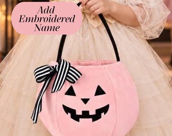 May include: A light pink pumpkin-shaped bucket with a black handle and a black and white striped bow. The bucket features a black jack-o'-lantern face with triangular eyes and a jagged mouth. A pink oval overlay displays the text Add Embroidered Name.