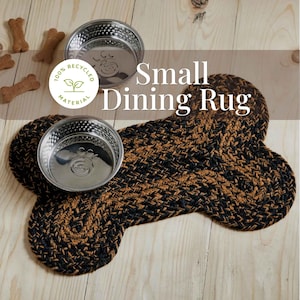 May include: A bone-shaped braided rug in black and brown with two stainless steel bowls placed on top. The rug features a circular label with the text 100% RECYCLED MATERIAL and the words Small Dining Rug are printed in white over the image.