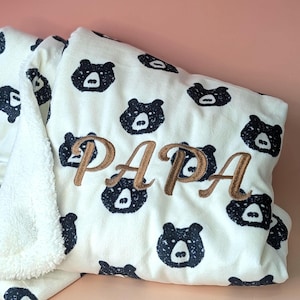 May include: A cream blanket featuring a navy bear face pattern and the embroidered word "PAPA" in brown. The blanket has a soft, plush texture. Ideal for a cosy and comfortable feel.