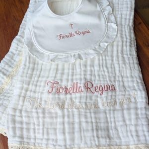 May include: A white muslin baby blanket and matching bib with ruffled edges. The bib features an embroidered cross and the name Fiorella Regina in pink. The blanket is embroidered with Fiorella Regina and The Lord bless and keep you.