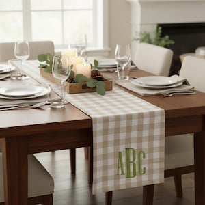 May include: A dining table set for a meal, featuring a beige and white checkered table runner with green embroidered initials. The table is set with white plates, glasses, candles, and a wooden centerpiece with greenery.