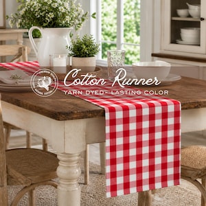 May include: A red and white checkered cotton table runner is draped over a wooden table. The runner has the words "Cotton Runner" and "Yarn Dyed - Lasting Color" printed on it. The table is set with plates, a white pitcher, and a small potted plant.