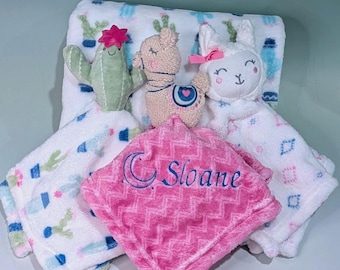 May include: A white blanket with a cactus print, a pink chevron-patterned blanket embroidered with the name Sloane and a crescent moon, a plush green cactus toy, a tan llama plush, and a white plush animal with a pink bow.