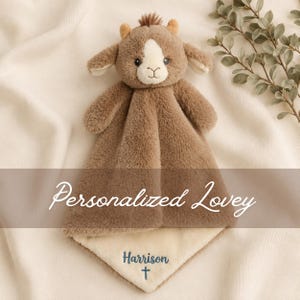Könnte beinhalten: Ein plüschiges, braunes Kuscheltier mit weicher, flauschiger Textur, bestehend aus einem kleinen Kopf mit Ohren und einem Haarbüschel. Die Decke ist auf der Unterseite cremefarben und mit dem Namen Harrison sowie einem kleinen Kreuz bestickt.