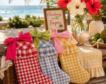 Puede incluir: Tres calcetines navideños con patrones de cuadros en rojo, azul y amarillo, cada uno con un puño con volantes y un gran lazo de tela. Un letrero enmarcado dice Christmas in the Tropics, junto a flores de hibisco y una piña.