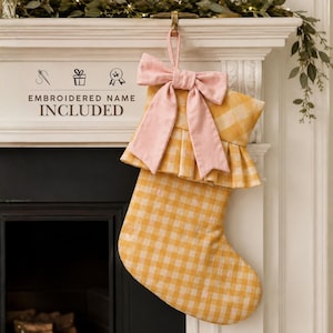 May include: A yellow and white gingham Christmas stocking featuring a large, solid light pink fabric bow at the top. The stocking has a ruffled cuff and includes the text EMBROIDERED NAME INCLUDED with icons of a needle, gift, and star.