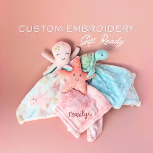 Könnte beinhalten: Drei Plüsch-Schmusetücher mit einem rosa Oktopus, einem korallenfarbenen Seestern und einer hellblauen Schildkröte. Das rosa Tuch ist mit dem Namen Marilyn bestickt. Der Text darüber lautet CUSTOM EMBROIDERY Gift Ready. Weiche Stofftexturen.