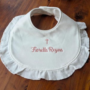 A white baby bib with a red embroidered name tag that says "Fiorella Regina" on a wooden surface.