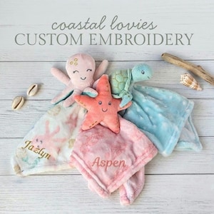 May include: Three plush baby security blankets featuring a pink octopus, a coral starfish, and a light green sea turtle. The blankets are embroidered with the names Jazlyn and Aspen. Text reads coastal lovies CUSTOM EMBROIDERY.