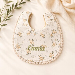 A white bib with a floral pattern and the name "Emma" embroidered on it, placed on a white surface next to some green leaves.