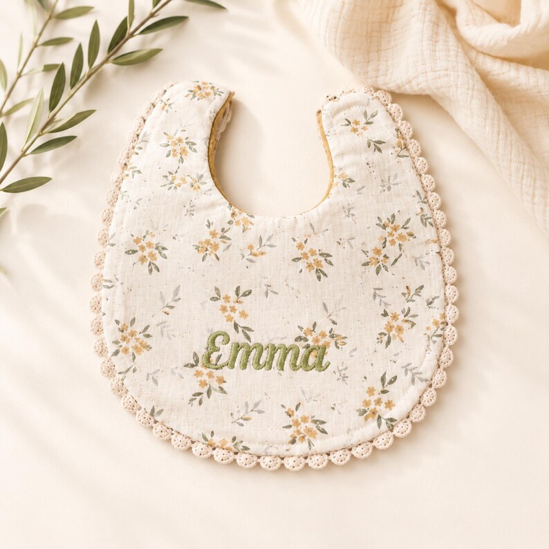 A white bib with a floral pattern and the name "Emma" embroidered on it, placed on a white surface next to some green leaves.