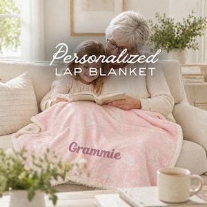 May include: A soft, light pink lap blanket with a white floral pattern and a cream-colored sherpa lining. The blanket features the embroidered name Grammie in a dark pink script font, draped over the laps of two individuals sitting together.