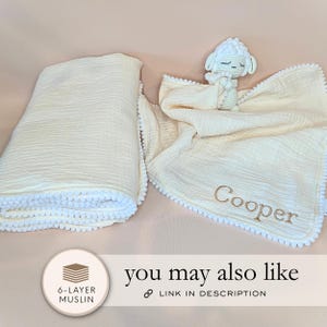a white blanket with a crocheted sheep toy and a personalized name tag, placed on a light pink background.