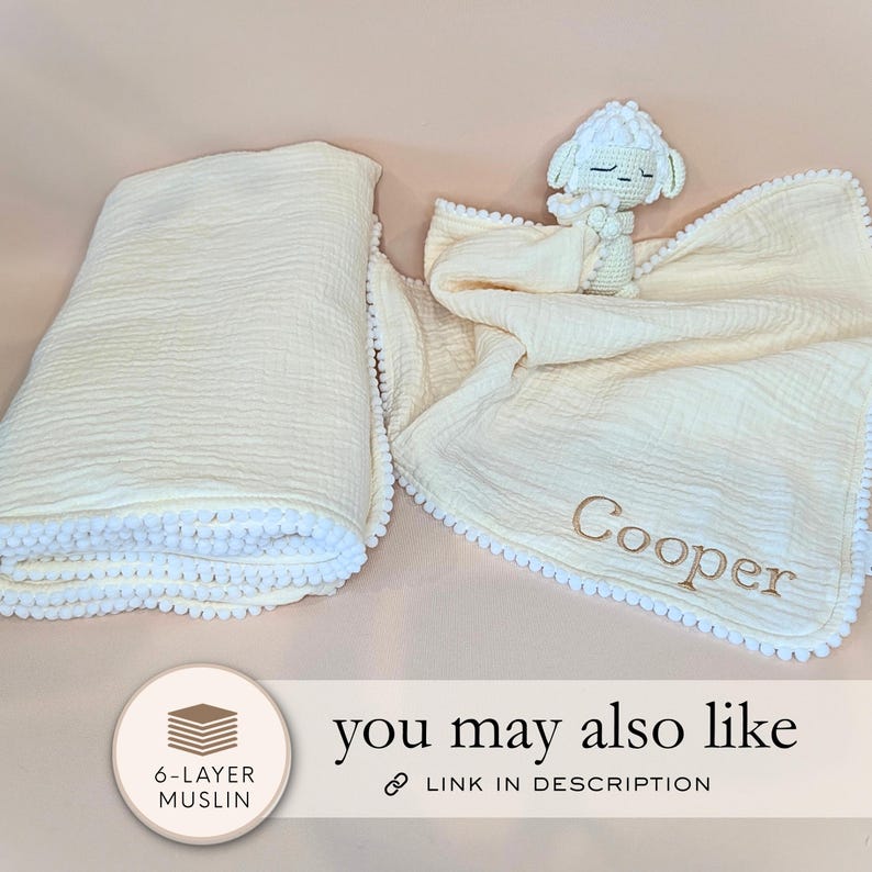 a white blanket with a crocheted sheep toy and a personalized name tag, placed on a light pink background.