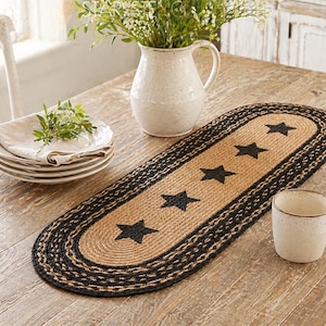 May include: Oval jute table runner with black stars and a black border. The runner is on a wooden table, with a stack of plates, a vase with flowers, and a mug.