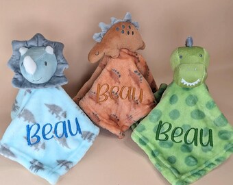 May include: Three plush dinosaur security blankets featuring soft fabric heads and square bodies. The blue blanket has a triceratops head, the orange one has a stegosaurus head, and the green one has a t-rex head. Each blanket is embroidered with the name Beau.