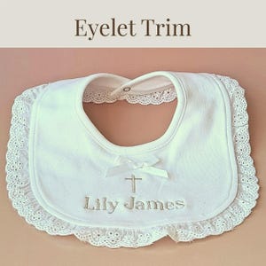 A white baby bib with a lace trim and a white ribbon with a cross on it, with the name "Lily James" embroidered on it.