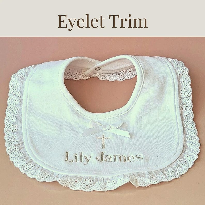 A white baby bib with a lace trim and a white ribbon with a cross on it, with the name "Lily James" embroidered on it.
