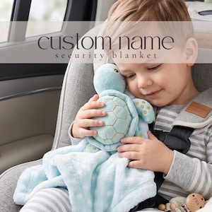May include: A light blue plush security blanket featuring a stuffed turtle head and shell with gold embroidered details. The soft fabric has a white polka dot pattern. The text custom name security blanket is overlaid on the image.