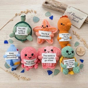 May include: Seven crocheted sea creatures including a green turtle, a pink starfish, an orange seahorse, a blue fish, a pink octopus, a pink seashell, and a green turtle, each holding a white card with encouraging text on a light wooden surface.