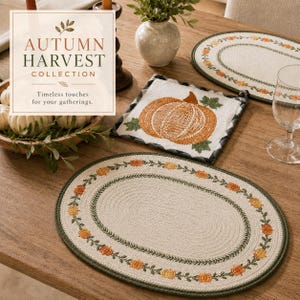 Op de afbeelding: Twee ovale gevlochten placemats met een crèmekleurig midden en een rand met oranje pompoenen en groene ranken, naast een vierkante pannenlap met een oranje pompoenafbeelding en de tekst AUTUMN HARVEST COLLECTION.
