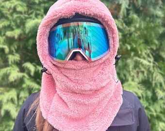 Könnte beinhalten: Eine rosafarbene Fleece-Balaclava mit Kapuze und einer blauen, verspiegelten Skibrille.