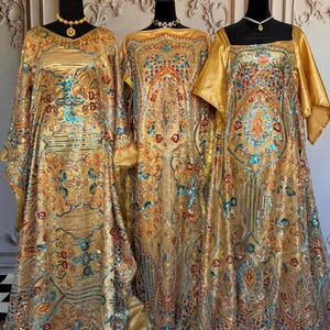 May include: Three gold sequined gowns with intricate floral patterns and colorful accents. The gowns are displayed on mannequins against a gold and white background.