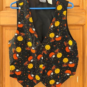 May include: A black vest featuring a repeating Halloween-themed pattern of orange pumpkins, yellow full moons, brown fences, and small white cats. The garment has a V-neckline, three front buttons, and a label reading Basic Editions at the collar.