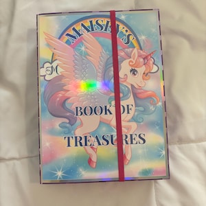 Op de afbeelding: Een rechthoekig boek met een eenhoornillustratie en de tekst "Maisey's Book of Treasures". De kaft heeft een regenboog, sterren en een roze elastiek. De eenhoorn heeft een roze manen en vleugels.