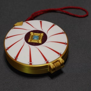 May include: A round, gold-colored trinket box with a white and red patterned lid. The lid has a small, square, blue gem in the center. The box has a red string attached to it.