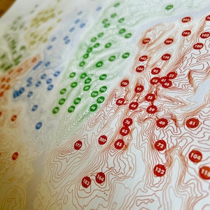 Puede incluir: Una impresi&oacute;n detallada de un mapa topogr&aacute;fico con l&iacute;neas de contorno en tonos naranja y marr&oacute;n. Puntos de colores en verde, azul y rojo est&aacute;n dispersos por el mapa, cada uno con un n&uacute;mero. El mapa est&aacute; impreso en papel color crema.