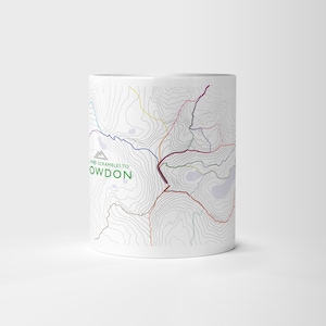 Puede incluir: Taza de cerámica blanca con un diseño de mapa topográfico que muestra líneas de contorno y senderos de senderismo coloridos. La taza tiene el texto "AND SCRAMBLES TO SNOWDON" en verde. Un icono de montaña está encima del texto.