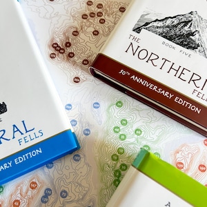 Puede incluir: Primer plano de tres libros con cubiertas blancas y lomos de colores, apoyados sobre un mapa. Los libros se titulan "The Northern Fells" y "A Pictorial Guide to the Lake District". El mapa presenta l&iacute;neas de contorno y puntos numerados.