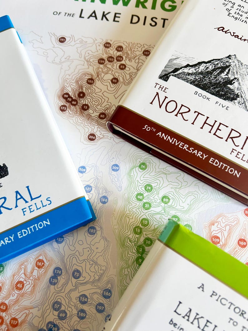 Puede incluir: Primer plano de tres libros con cubiertas blancas y lomos de colores, apoyados sobre un mapa. Los libros se titulan "The Northern Fells" y "A Pictorial Guide to the Lake District". El mapa presenta l&iacute;neas de contorno y puntos numerados.