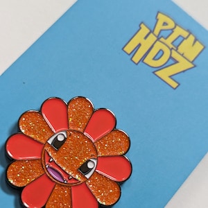 May include: Orange glitter enamel pin with a red flower design and a cartoon face with fangs. The pin says "PIN HDZ" in yellow letters.