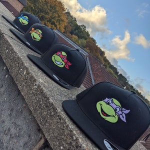 Pode incluir: Quatro bonés de beisebol pretos com diferentes personagens das Tartarugas Ninja Mutantes Adolescentes bordados na frente. Os personagens são Leonardo, Rafael, Donatello e Michelangelo.