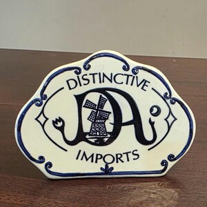 May include: Cream-colored ceramic plaque with a blue border and text. The plaque reads "DISTINCTIVE DA IMPORTS" in black lettering. The "DA" features a windmill design. The border has decorative swirls and diamond shapes.