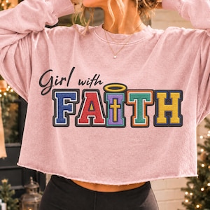 May include: A light pink long-sleeve sweatshirt with a raw-edge hem. The front features the black cursive text Girl with and the block-lettered word FAITH in blue, red, purple, green, and yellow, with a gold cross and halo design.