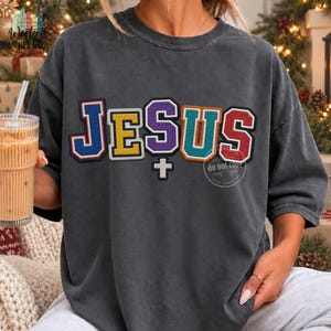 May include: A charcoal gray short-sleeve t-shirt featuring the word JESUS in large, multi-colored varsity-style letters with a white cross symbol underneath. Each letter has a distinct color including blue, yellow, purple, teal, and red.