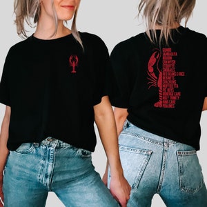 May include: Black t-shirt with a red crawfish graphic on the front and a list of Cajun food items on the back. The text includes: Gumbo, Jambalaya, Bougin, Crawfish, Etouffee, Snowballs, Red Beans & Rice, Beignets, Cracklins, Muffuletta, Po Boys, Odberge Cake, Holy Trinity, King Cake, Pralines.