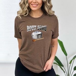 May include: Brown t-shirt with the text "ROUX GURU" in silver lettering. A silver pot with a wooden spoon is pictured below the text.