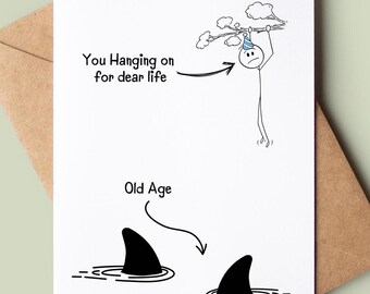 Peut inclure: Une carte d'anniversaire humoristique avec une illustration de bonhomme bâton. Le bonhomme bâton est accroché à une branche avec le texte "You Hanging on for dear life". Sous le bonhomme bâton, il y a deux nageoires de requin avec le texte "Old Age".