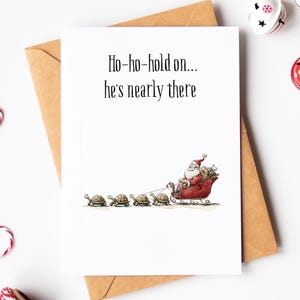 Puede incluir: Tarjeta de Navidad con una ilustración humorística de Papá Noel en un trineo rojo tirado por cuatro tortugas. La tarjeta tiene el texto "Ho-ho-hold on... he's nearly there" sobre un fondo blanco, con un sobre marrón.
