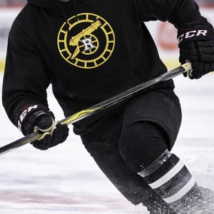 May include: Black hoodie with a yellow and black circular logo featuring a cannon and the letter "R". The person is wearing black hockey gloves and black trousers with white and black striped socks. A hockey stick is visible.