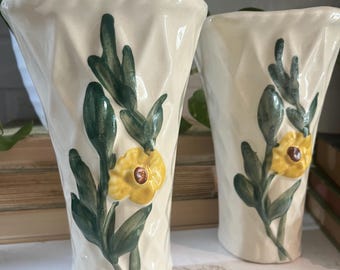 May include: Two off-white ceramic vases with a textured surface. Each vase features a hand-painted design of a yellow flower with a brown center and green leaves. The vases are displayed on a white surface.