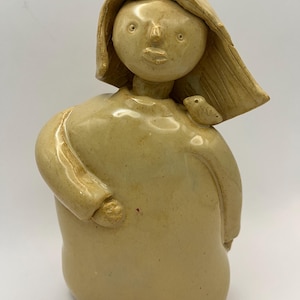 May include: A ceramic figurine of a person with long hair and a bird on their shoulder. The figurine is painted in a light brown color.