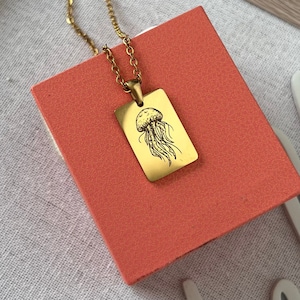 May include: Gold necklace with a rectangular pendant featuring a detailed line drawing of a jellyfish.
